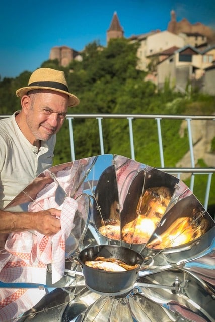 Alain présente le SUNplicity, appareil de cuisson solaire, médaille d'or au concours Lépine. Chapeau et torchon pour soulever le couvercle de la cocotte sont de mise par cette radieuse journée ensoleillée. En fond, on distingue les hauteurs d'un village du Tarn. Le SUNplicity, le four solaire français par excellence.